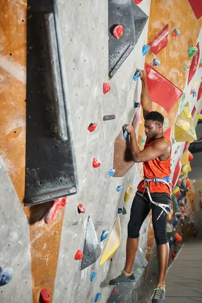 Tiro vertical del hombre americano africano deportivo en camisa naranja colgando de sus dedos en la pared de escalada - foto de stock