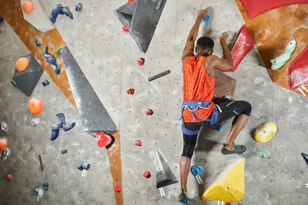 Young african american man with alpine harness ascending up climbing wall, back view, bouldering — Stock Photo