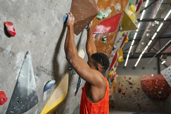 Young african american man with alpine harness hanging on huge boulder on climbing rock wall — Stock Photo