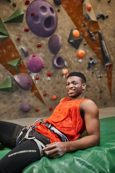 Plano vertical de alegre afroamericano relajante en almohadilla de bouldering y sonriendo alegremente a la cámara - foto de stock