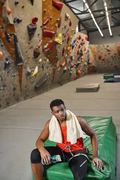 Hombre americano africano deportivo que se relaja en la almohadilla de choque después de escalar pared de roca, toalla y botella de agua - foto de stock