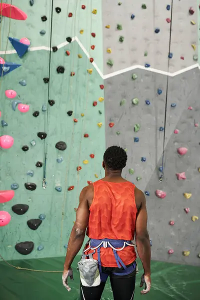 Vertical shot of sporty african american man in orange shirt with alpine harness near rock wall — Stock Photo