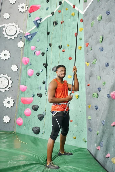 Tiro vertical del hombre afroamericano en camisa naranja usando cuerda trepadora en la pared de roca, bouldering - foto de stock