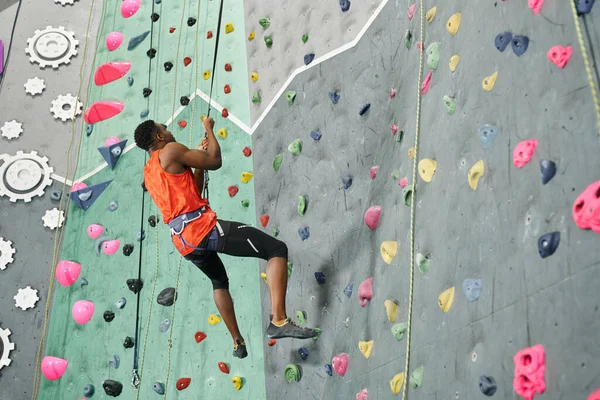 Athletic african american man in orange shirt hanging on his climbing rope and looking up, rock wall — Stock Photo