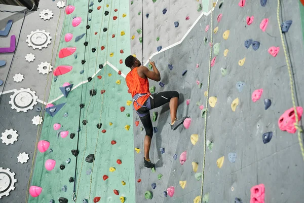 Young african american man in orange shirt climbing up rock wall with rope and alpine harness — Stock Photo