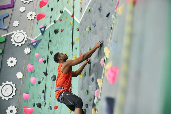 Young african american man climbing up bouldering wall with safety rope and alpine harness — Stock Photo