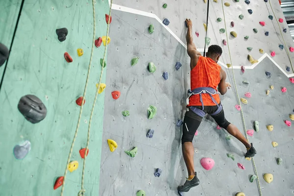 Back view of young african american man in orange shirt climbing up bouldering wall with safety rope — Stock Photo