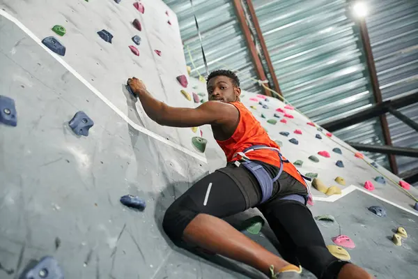 Atletico uomo africano americano con corda di sicurezza arrampicata su parete rocciosa e guardando verso il basso la fotocamera — Foto stock