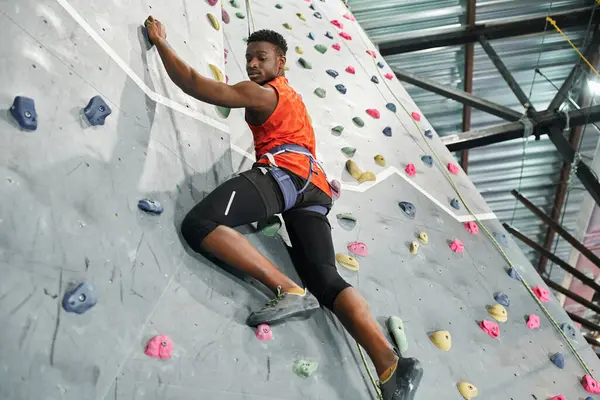 Homme afro-américain fort en chemise orange escalade mur avec corde de sécurité et regardant vers le bas — Photo de stock