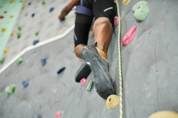 Cropped view of legs of sporty african american man climbing up rock wall with safety rope — Stock Photo