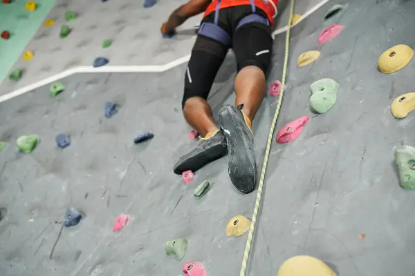 Vista cortada das pernas do homem americano africano forte escalando acima da parede bouldering com arnês alpino — Fotografia de Stock