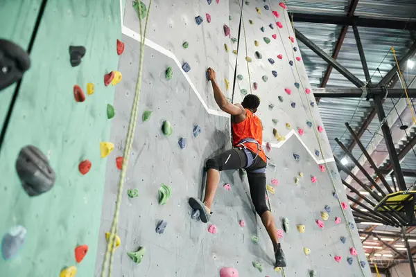 Sporty african american man in orange shirt climbing up bouldering wall gripping strongly on rocks — Stock Photo