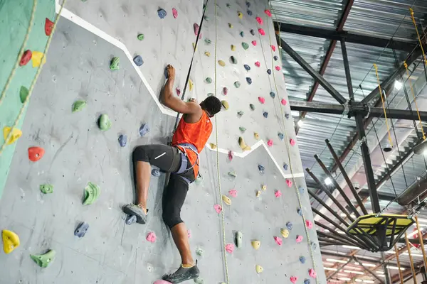 Strong african american man in orange shirt using safety rope and alpine harness to climb up wall — Stock Photo