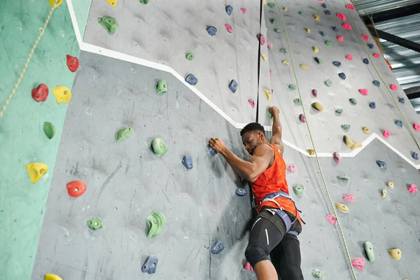 Desportivo jovem afro-americano homem usando corda de segurança e arnês para subir a parede de rocha, bouldering — Fotografia de Stock