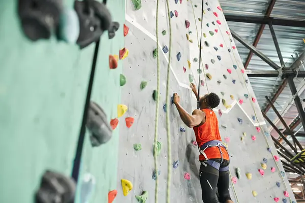 Forte giovane afroamericano in camicia arancione con imbracatura alpina arrampicata su parete rocciosa — Foto stock