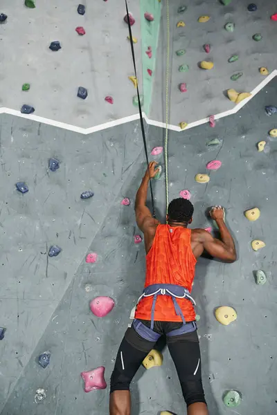 Forte giovane afroamericano con imbracatura alpina e corda di sicurezza arrampicata su parete, bouldering — Foto stock
