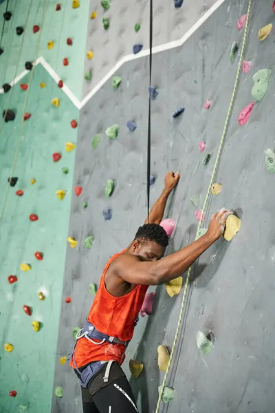 Красивый африканский американец в оранжевой рубашке с альпийской аркадой, взбирающийся на валунную стену — стоковое фото