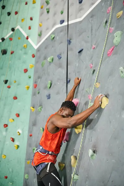 Hombre afroamericano guapo en camisa naranja con arnés alpino trepando por la pared de bouldering - foto de stock