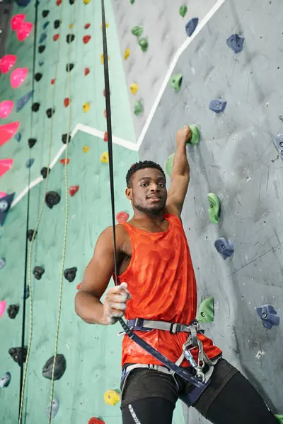 Guapo afroamericano hombre posando mientras ascendente hasta bouldering pared usando cuerda de seguridad, deporte - foto de stock