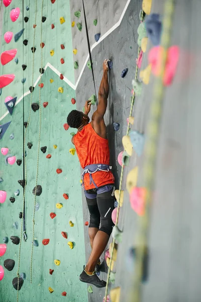 Sporty african american man in orange shirt with alpine harness and safety rope climbing up wall — Stock Photo