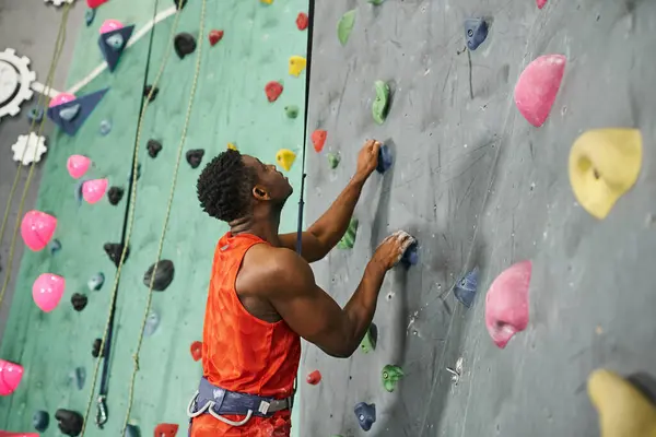 Guapo joven afroamericano hombre de camisa naranja mirando hacia arriba mientras trepa por la pared de bouldering - foto de stock