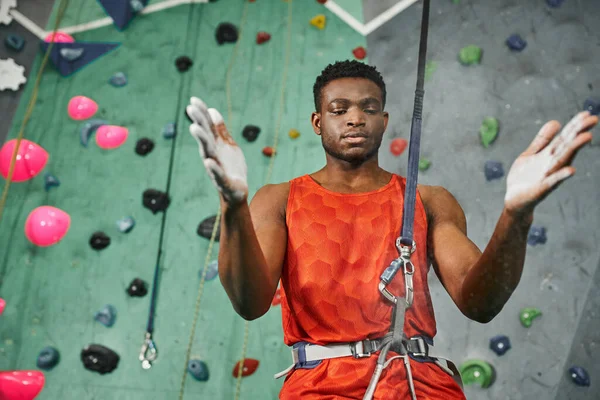 Молодой атлетичный африканский американец в оранжевой рубашке с канатом безопасности с использованием талькового порошка, боулдеринг — стоковое фото
