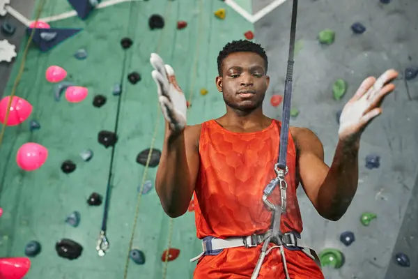 Hombre afroamericano bien parecido en camisa naranja con cuerda de seguridad usando tiza de gimnasio, bouldering - foto de stock