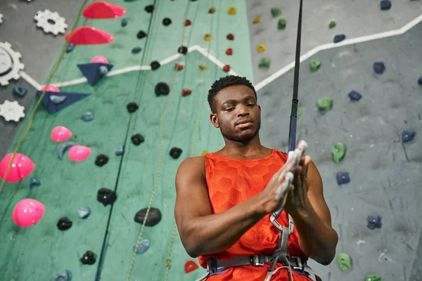 Homem americano africano bonito na camisa alaranjada posando ao lado da parede bouldering usando o pó do talco — Fotografia de Stock