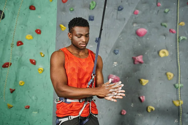 Joven atlético afroamericano hombre en camisa naranja con cuerda de seguridad usando talco en polvo, bouldering - foto de stock