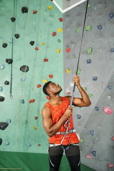 Tiro vertical del joven afroamericano en camisa naranja mirando hacia arriba antes de subir por la pared de roca - foto de stock