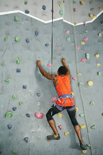 Back view of african american man in orange shirt climbing actively up rock wall, bouldering concept — Stock Photo