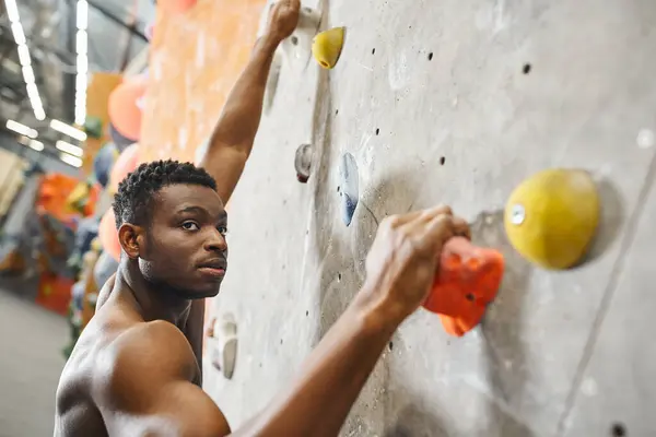 Handsome fit african american man with his shirt off gripping on boulders while climbing up wall — Stock Photo