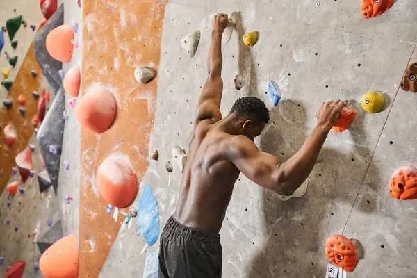 Sporty african american man with his shirt off gripping on rocks while climbing up bouldering wall — Stock Photo