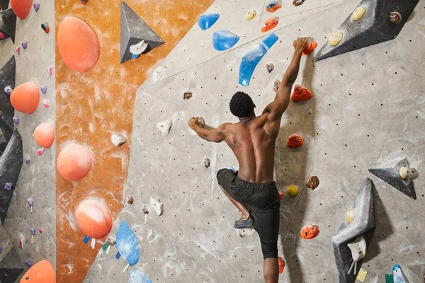 Muscoloso uomo afroamericano senza maglietta che afferra fortemente i massi mentre si arrampica sulla parete rocciosa — Foto stock