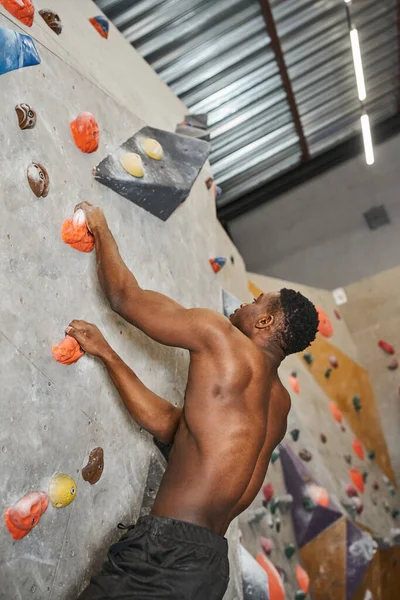 Good looking topless african american man gripping on rocks while climbing up bouldering wall — Stock Photo