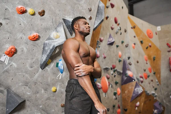 Uomo africano americano in topless sportivo in pantaloni neri in posa accanto al muro di bouldering e guardando altrove — Foto stock