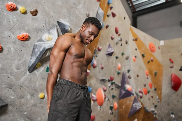Gioioso muscolare africano americano uomo in pantaloni neri in posa topless accanto al muro di bouldering — Foto stock