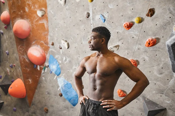 Hombre afroamericano sin camisa de buen aspecto junto a la pared de escalada con las manos akimbo, mirando hacia otro lado - foto de stock
