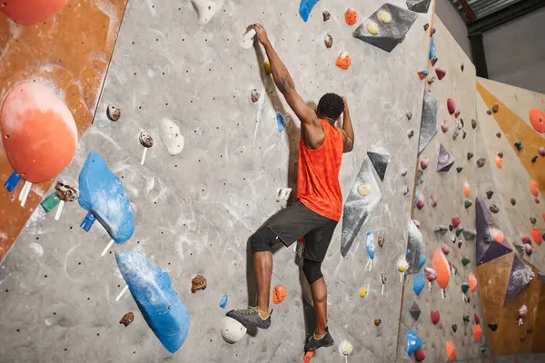 Athletic african american man in orange shirt gripping on boulders while climbing up rock wall — Stock Photo