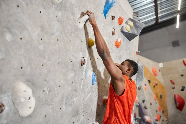 Young african american man in orange shirt gripping on rocks while climbing up wall, bouldering — Stock Photo