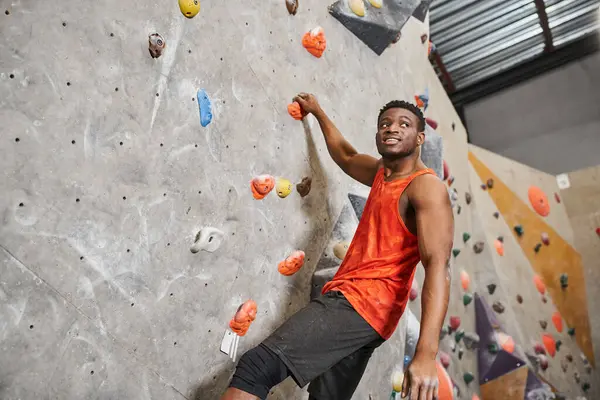 Alegre afroamericano hombre en naranja camisa ascendiendo hasta pared de roca y sonriente mirando hacia otro lado - foto de stock