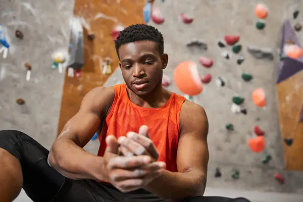 Hombre afroamericano guapo en camisa naranja mirando las manos en tiza gimnasio con fondo de pared de roca - foto de stock