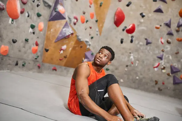Sportif jeune homme afro-américain en chemise orange assis à côté du mur de blocs et regardant loin — Photo de stock