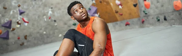 Homme afro-américain athlétique en chemise orange assis à côté de la paroi rocheuse et regardant ailleurs, bannière — Photo de stock