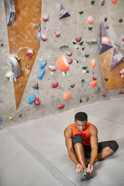 Good looking african american man sitting on floor and tying his shoes next to climbing rock wall — Stock Photo