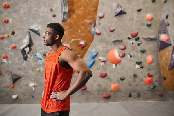 Bonito afro-americano homem posando em perfil com as mãos em quadris ao lado de escalada parede de rocha — Fotografia de Stock