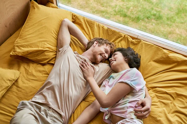 Vista dall'alto di uomo felice abbracciare ragazza asiatica e sdraiati insieme su biancheria da letto gialla a casa, coppia — Foto stock