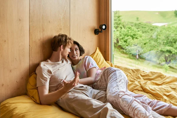 Redhead man talking to cheerful asian girlfriend while lying together in bed next to window — Stock Photo
