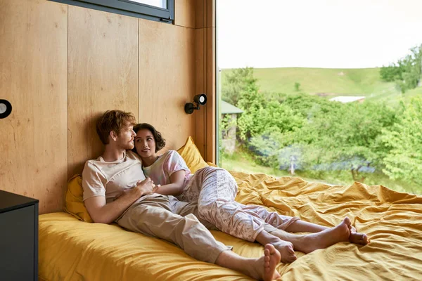 Redhead man lying on bed with cheerful asian girlfriend at looking at window with natural view — Stock Photo