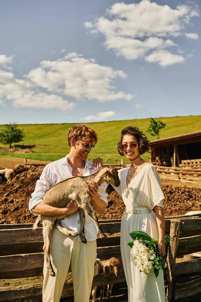 Newlyweds in countryside, happy man holding goat near asian bride in white dress, rustic wedding — Stock Photo